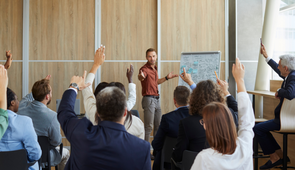 Un giovane insegnante davanti a lavagna a fogli mobili e un gruppo di allievi con la mano alzata per rispondere